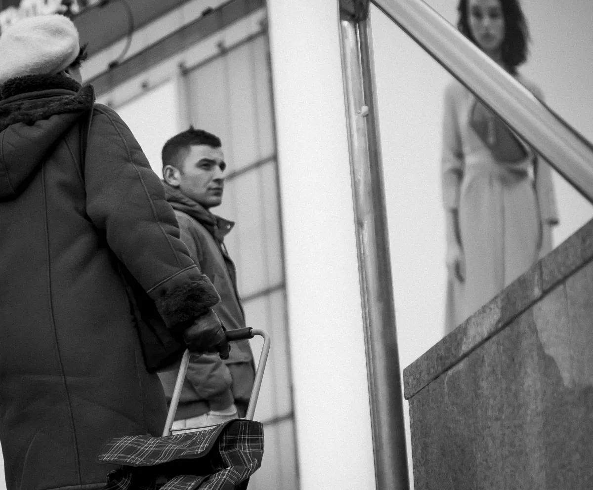 Older woman with a shopping trolley crossing in front of a young man and a blurred female poster